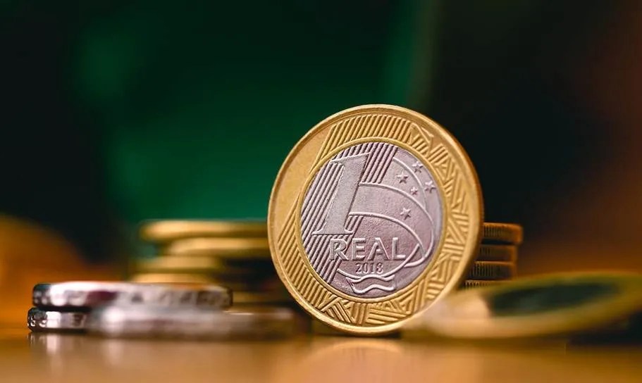 Moeda de 1 real em destaque sobre a mesa, com outras moedas desfocadas ao fundo.