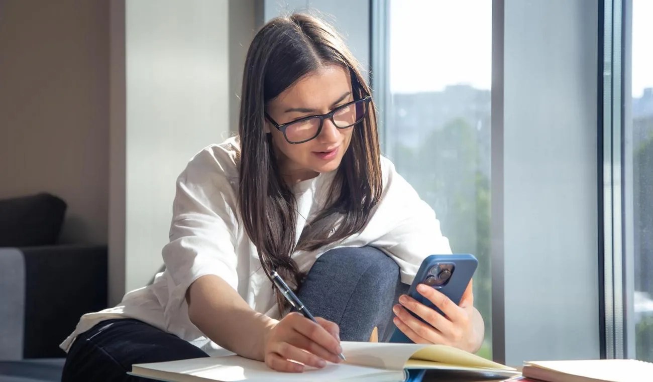 Mulher de óculos fazendo anotações em um caderno enquanto consulta o celular, avaliando custo de oportunidade dos investimentos.