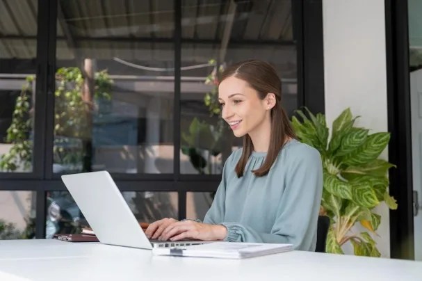 Mulher sorrindo enquanto usa notebook em uma mesa de escritório com papéis e plantas ao fundo, pesquisando sobre produtos do mercado financeiro.