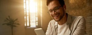 Homem em um ambiente interno bem iluminado, usando fones e olhando o celular enquanto faz anotações em um caderno ao lado do teclado, pesquisando investimentos sem imposto de renda.
