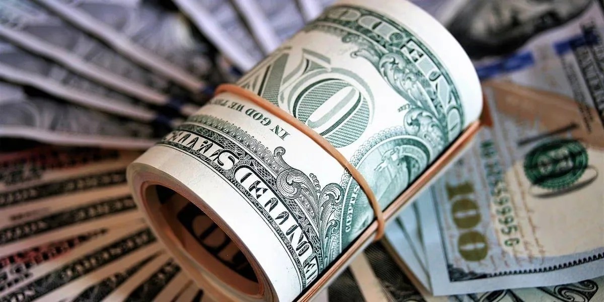 Rolo de notas de dólar cercado por cédulas americanas espalhadas, representando o dólar como ativo de proteção patrimonial e reserva de valor no mercado financeiro.