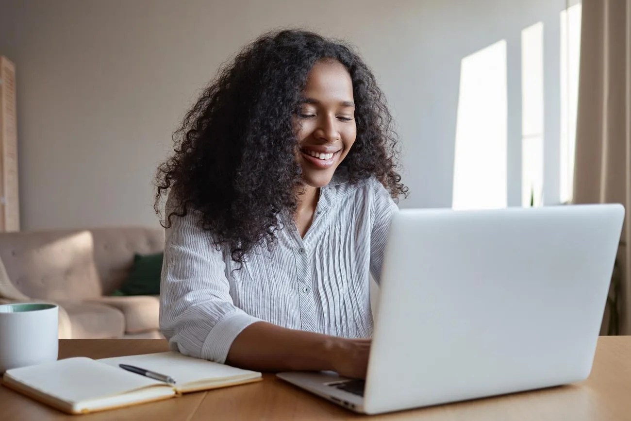Mulher sorrindo enquanto usa notebook em uma mesa com caderno, caneta e caneca, pesquisando como escolher uma boa reserva de valor.