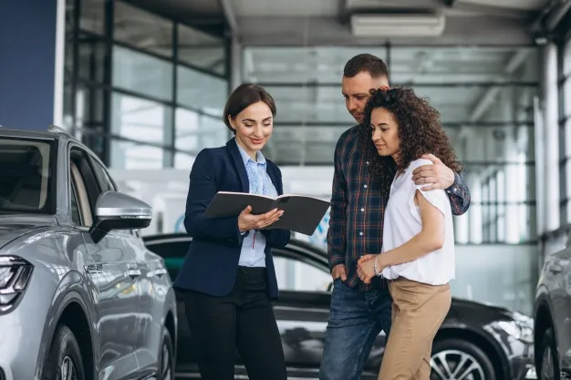 Fotografia de casal conversando com uma corretora de automóveis. Ela está com uma pasta na mão e no mundo tem alguns HUV.