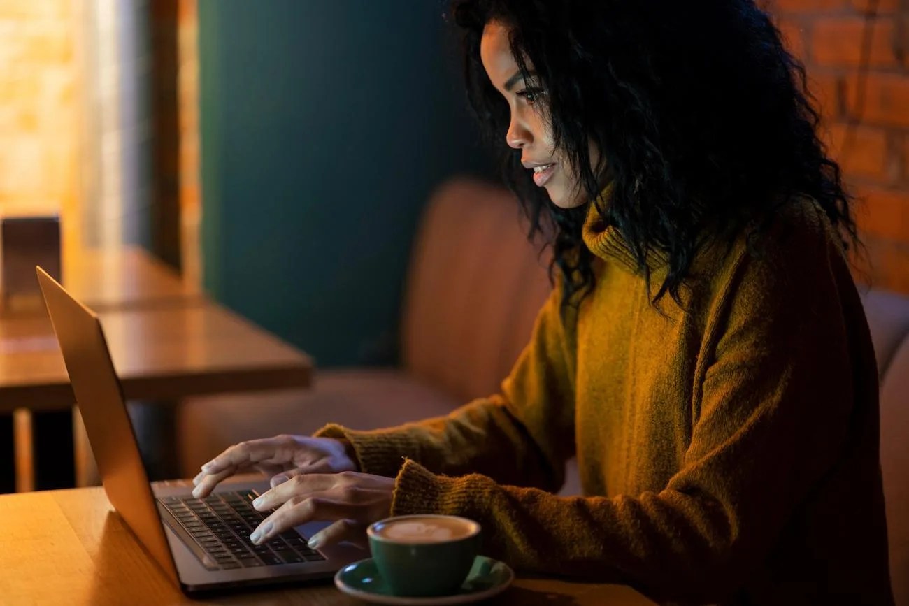 Mulher sentada em uma cafeteria, usando um laptop e analisando informações financeiras relacionadas a indicadores de liquidez, com uma xícara de café sobre a mesa.