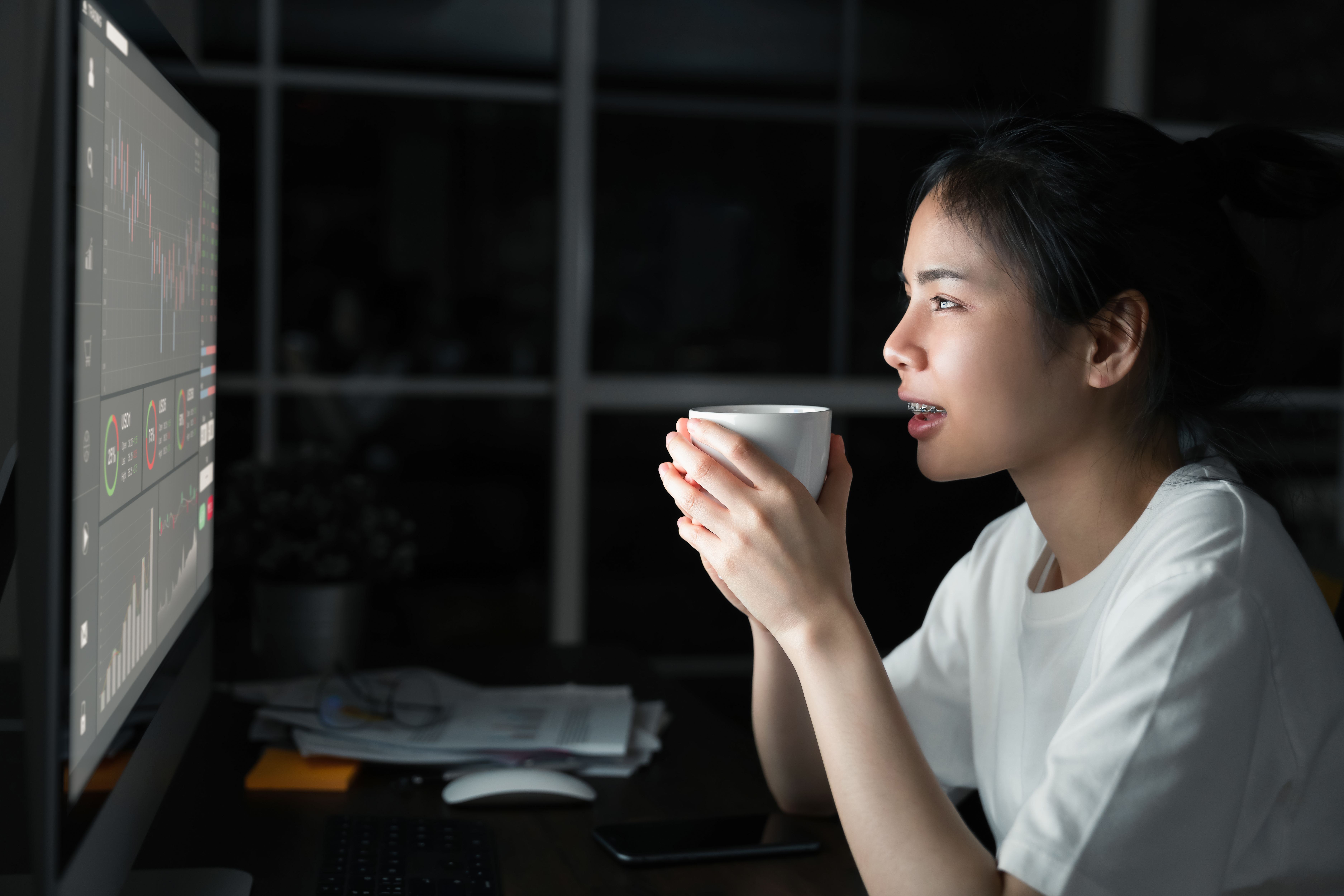 Fotografia de mulher analisando seu home broker. Ela está segurando uma xícara, já a luz está apagada.