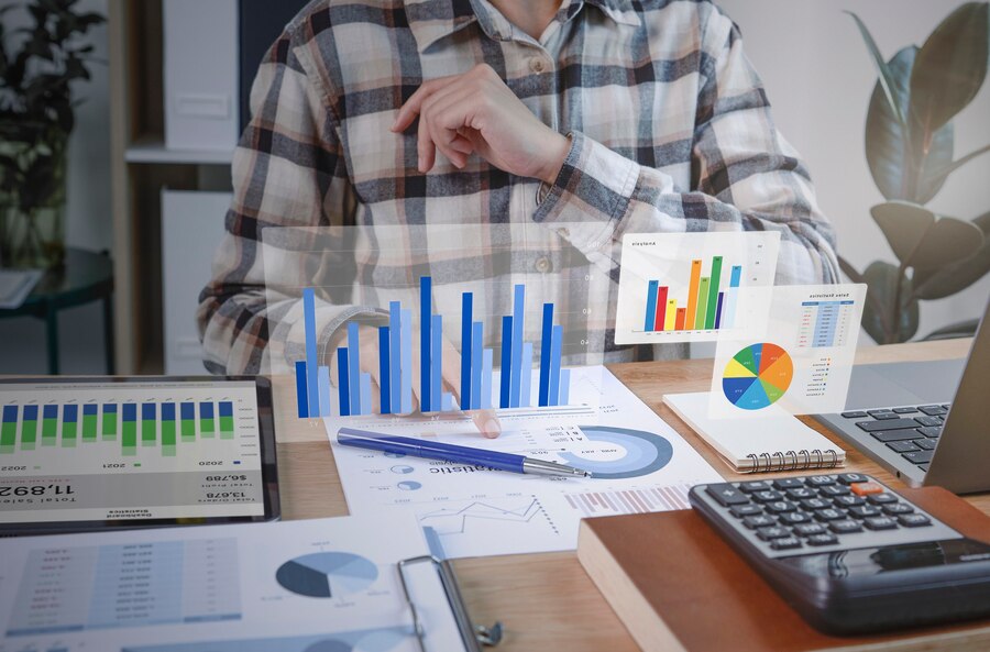 Fotografia de homem analisando gráficos da especulação financeira. Na mesa há vários gráficos, calculadora e notebook