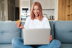 Fotografia de mulher sentada no sofá da sala com o notebook no colo. Ela está com um cartão na mão, provavelmente assinando o Expert Pass XP.