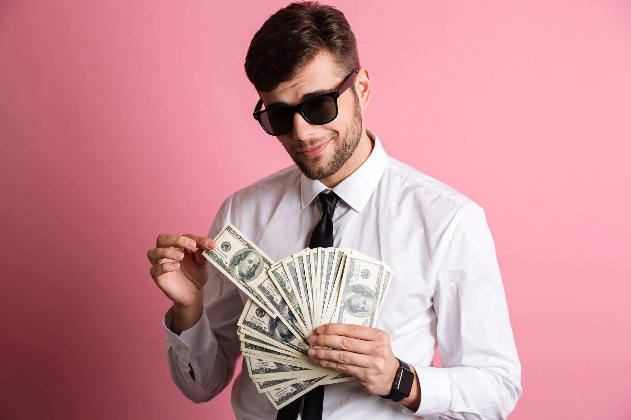 Fotografia de homem com um leque de cédulas de dólar nas mãos. Ele usa camisete, gravata e óculos escuros.