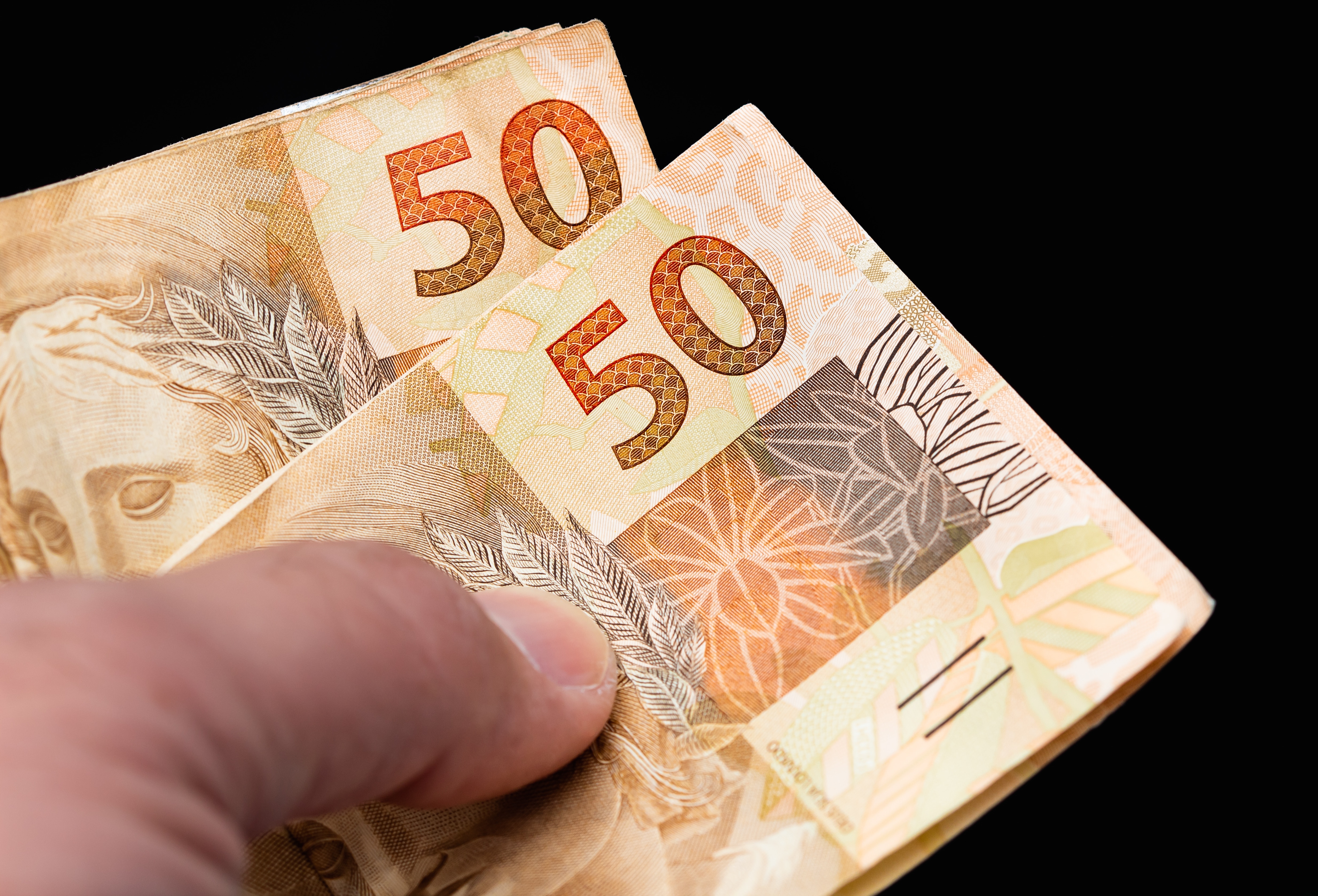Fotografia de uma mão segurando duas notas de R$50,00.