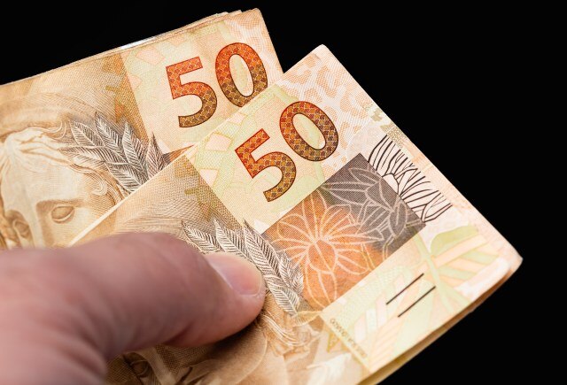Fotografia de uma mão segurando duas notas de R$50,00.