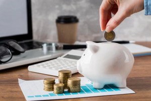 Pessoa colocando uma moeda em um cofrinho sobre a mesa, com pilhas de moedas, calculadora, notebook e gráfico de organização financeira, planejando como fazer uma reserva de emergência.