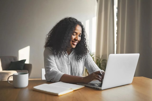 Mulher sorrindo enquanto usa notebook em uma mesa com caderno aberto e caneca, pesquisando dicas para fazer uma boa reserva de emergência.