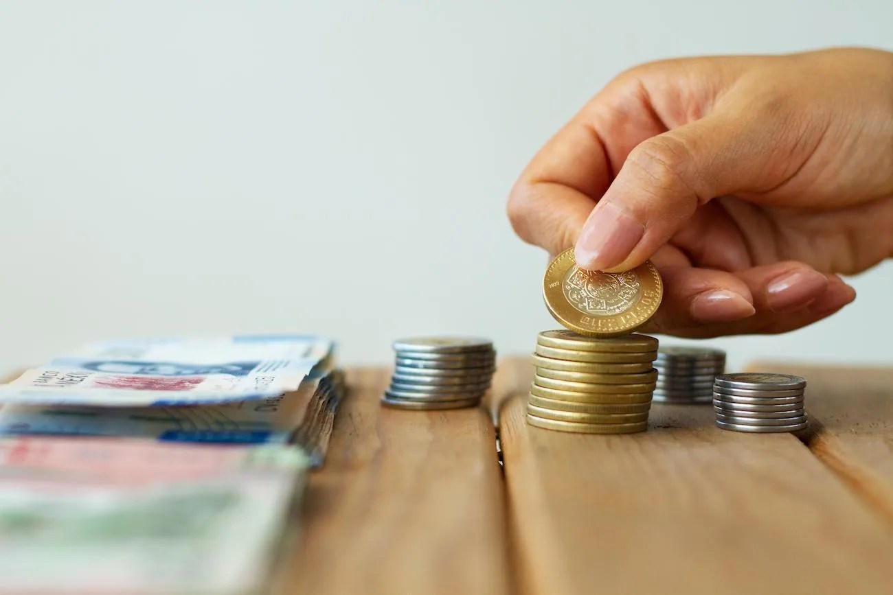 Pessoa colocando uma moeda sobre pilhas de moedas ao lado de cédulas em uma mesa de madeira, representando planejamento financeiro para viver de renda extra.