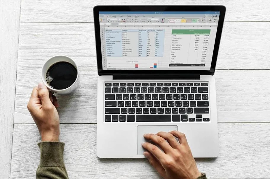 Pessoa usando notebook com uma planilha no excel aberta, segurando uma xícara de café sobre uma mesa branca de madeira, em busca da melhor planilha de gastos para organizar seu orçamento.