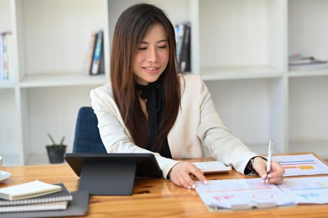 Mulher estudando sobre o mercado financeiro. Em sua mesa há alguns papeis com gráficos e tabelas, um tablet e alguns blocos de nota.