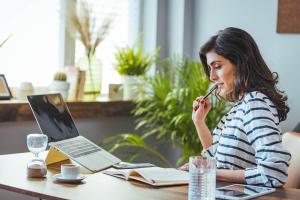 Mulher sentada à mesa de trabalho, em um ambiente claro e decorado com plantas, segura uma caneta entre os dedos e o rosto enquanto lê um caderno aberto refletindo sobre o melhor investimento de renda fixa. À sua frente, há um laptop elevado em suporte, um smartphone, um tablet, um copo de água e uma xícara de café sobre o pires e uma ampulheta.