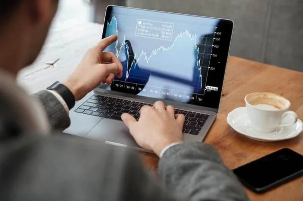 Pessoa acompanhando gráficos de movimento de mercado de um ativo em um notebook, com café e celular sobre a mesa, analisando a volatilidade para entender como investir melhor.