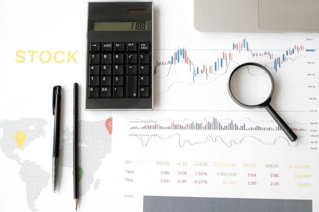 Calculadora e lupa sobre relatório financeiro de volatilidade nos investimentos em gráficos de preços com altas e quedas. Ao lado, há canetas, um mapa e parte de um notebook.